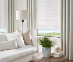 Custom draperies over fabric roman shades in a living room with white neutral and wooden tones. The window shades are partially open exposing a view of the ocean
