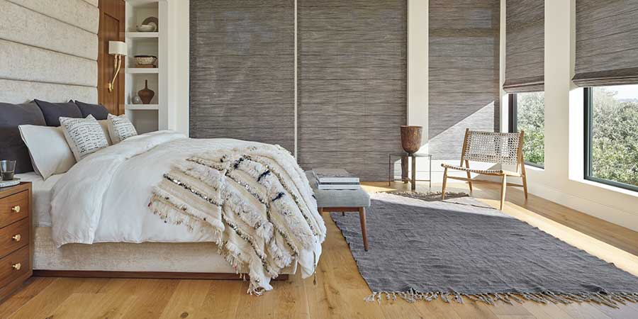 A neutral bedroom with alustra woven texture shades cascaded on two walls of windows in a neutral bedroom